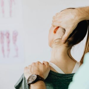Back view of crop faceless bearded orthopedist in wristwatch checking up neck of anonymous lady while standing in front of wall with paper drawings representing body anatomy
